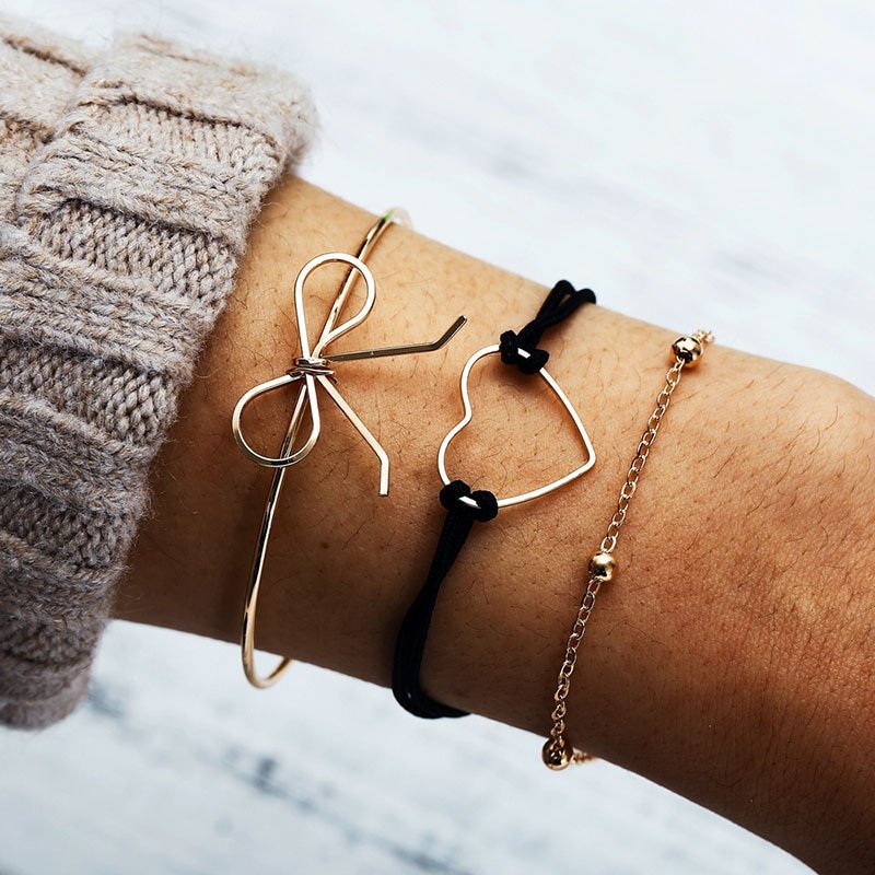 A person's wrist adorned with three bracelets including a Bohemian Rope Chain Bracelet for Women, a black cord bracelet with hearts, and a delicate chain bracelet, epitomizing the latest fashion trends.