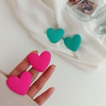 A hand holding two Colorful Heart-Shaped Acrylic Earrings for Women, with two green heart clips and a white cloth background, showcasing a trendy fashion accessory.