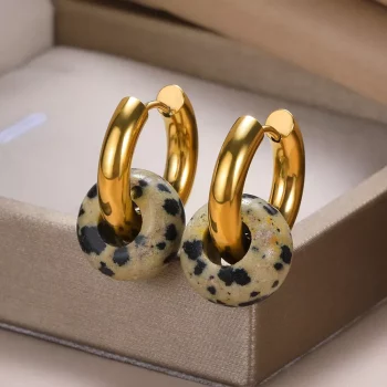 A pair of Women's Natural Stone Hoop Earrings with gold clasps and speckled beige and black ceramic rings, displayed in an open beige jewelry box, capture the essence of new fashion trends.